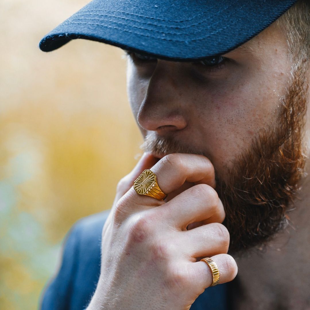 a man with a beard wearing a dark navy baseball cap, looking pensive. He is holding his chin with one hand, displaying rings with intricate designs on his fingers.